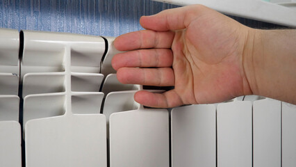 Unrecognizable man's hand check for heat and touch central heating system. White metal heating iron household radiator inside room. Budget in crisis concept. House in winter