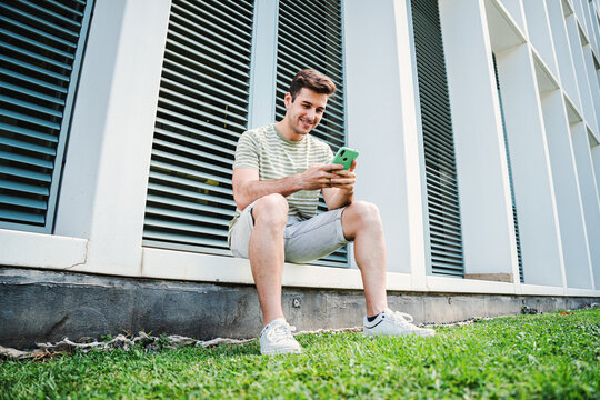 Caucasian teenage stundent enjoying using a mobile phone to watch the social media. Young guy with friendly expression smiling and having fun playing a video game with his cellphone sitting outside