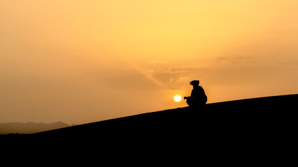 Silhouette of unidentified Berber man sitting, watching sunset on sand dunes at beautiful sunset in Sahara Desert, Morocco - obrazy, fototapety, plakaty