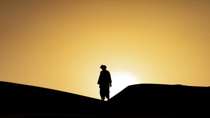 Silhouette of unidentified Berber man in his traditional clothing, a long robe on sand dunes at beautiful sunset in Sahara Desert, Morocco - obrazy, fototapety, plakaty
