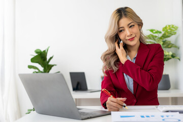 Attractive Asian female accountant talking on the phone with her business client while working with laptop and financial documents in the office