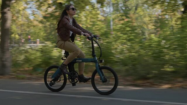 Stylish young woman riding an electric bicycle in city centre in the park during fall season. Concept of cycling sport activity and eco transportation by bike