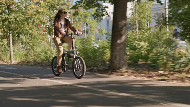 Stylish young woman riding an electric bicycle in city centre in the park during fall season. Concept of cycling sport activity and eco transportation by bike. 4k slow motion with copy space 