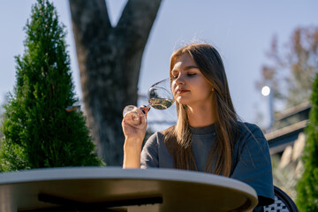 Fototapeta premium An adult girl sits at a table in the garden with a glass of wine and tastes a drink in the garden at winery