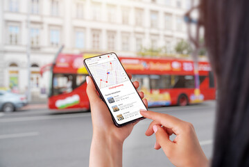 City Sightseeing app on smartphone in woman hands. City tourist bus in background - obrazy, fototapety, plakaty