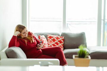 Smiling cute young woman sitting on sofa in casual red clothes in living room holding mobile phone
