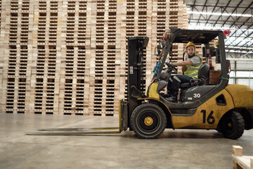 Warehouse worker wears safety helmet driving forklift truck in pallet factory. Skilled male...