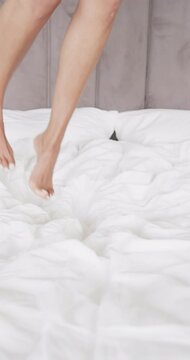 Vertical video of close up of legs of biracial woman jumping on bed at home, slow motion
