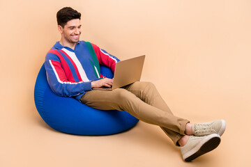 Full body length photo of cheerful businessman using computer when lying comfortable pouf at coworking isolated on beige color background