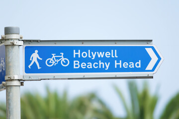Fototapeta premium A signpost showing a walkway and cycle path to Holywell and Beachy Head, shot against a blue summer sky and green foliage