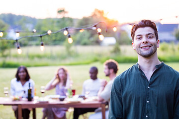 An attractive South European man with short brown hair in a green shirt laughs joyously with a wine glass in hand, as friends share the mirth in a garden barbecue setting, under the warm sunny skies