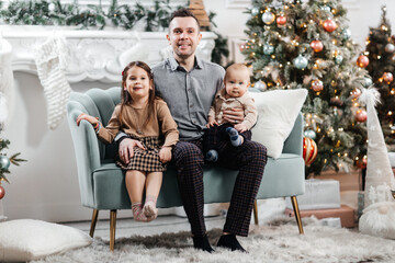 Dad spends time with his little children in front of a Christmas tree and Christmas decorations. Happy father and his son and daughter play together. Dad hugs his happy children