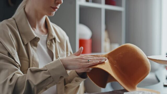 Tilting up shot of female millinery worker sitting in her craft store coating new orange hat with felt stiffener