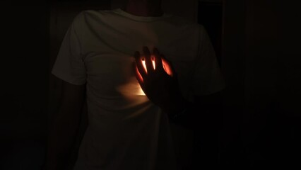 A man in a white T-shirt in a dark room holds his hand on his heart. Flashing light in rhythm with your heartbeat. Signs of stroke and heart disease.