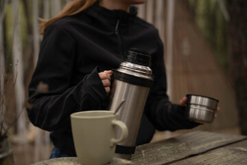 Outside a Glamping , Young girl uses the thermos of coffee on a wooden table outdoors during autumn.