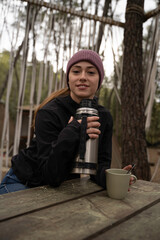 outside a Glamping , The smiling girl uses the thermos of coffee on a wooden table outdoors during...