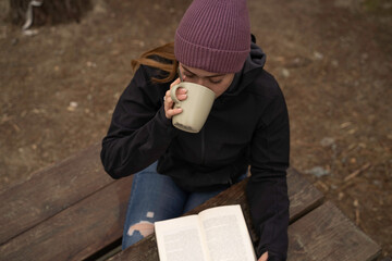 Fototapeta premium outside a Glamping , zenithal shot Smiling girl drinks coffee while reading a book on a wooden table outdoors during autumn.