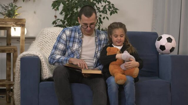 Happy family. A girl with toys listens to dad story. Father reads book to his daughter. Go to sleep. Family training in quarantine. Homeschooled girl. Dad reads a book to his daughter before bedtime