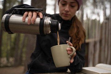 Fototapeta premium The girl wearing a winter hat pours coffee from her cup into the thermos and is sitting on a wooden table