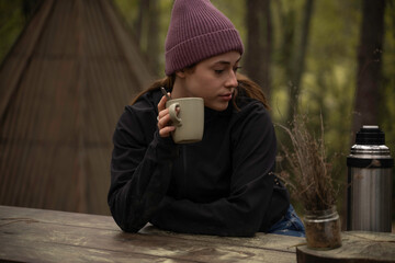 Glamping wooden teepee, a girl in autumn clothes is sitting outdoors on a wooden table while...