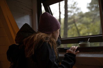 Girl in winter hat is looking out the window of a log cabin while drinking coffee from the cup.