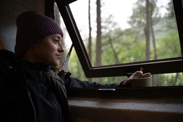 Girl in winter hat is looking out the window of a log cabin while drinking coffee from the cup.