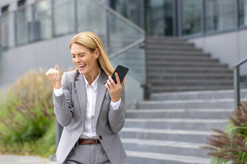 Fototapeta premium Successful mature joyful businesswoman received online notification message with good results achievement on phone, woman boss uses app on smartphone, walks outside office building in business suit