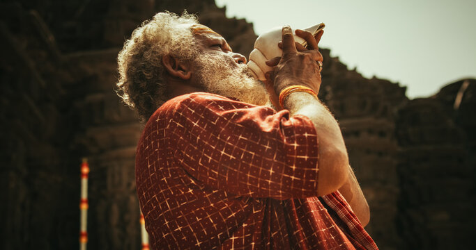 Hinduism, South Asian Religion and Cultural Footage: Authentic Shot of An Indian Senior Monk Blowing a Conch Shell in an Ancient Stone Temple. Elderly Man Performing Sacred Ritual on a Worship Day