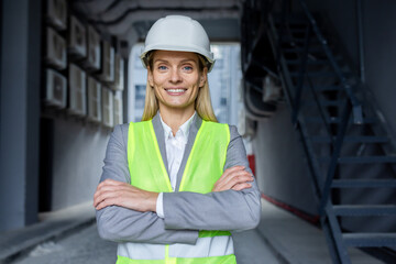 Portrait of mature successful female engineer, worker in hard hat and reflective vest with crossed arms smiling and looking at camera