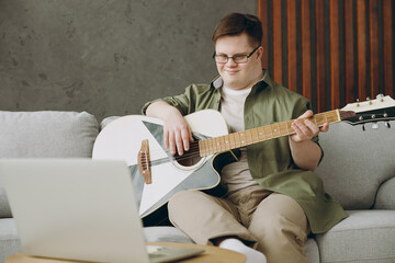 Young man with down syndrome wearing glasses casual clothes learn how to play guitar sitting on grey sofa couch stay at home flat rest spend free time in living room Genetic disease world day concept