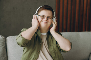 Young man with down syndrome wear glasses casual clothes listen to music in headphones sits on grey...