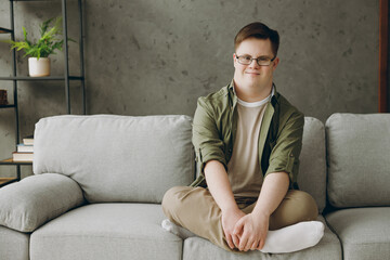 Full body young smiling man with down syndrome wear glasses casual clothes look camera sits on grey...