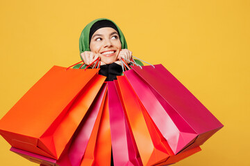Young smiling cheerful arab asian muslim woman wears green hijab abaya clothes hold shopping package bags look aside isolated on plain yellow background . Black Friday sale buy day, uae islam concept.