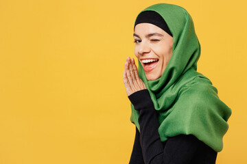 Young asian muslim woman wear green hijab abaya black clothes whispering gossip and tells secret behind her hand isolated on plain yellow background. People uae middle eastern islam religious concept.
