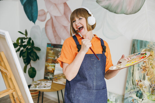 Elderly artist woman 50 years old in casual clothes stand near easel with artwork paint listen music in headphones sing song in microphone spend free time in living room indoor Leisure hobby concept