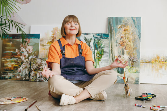 Full body elderly artist woman 50 years old wears casual clothes stand near easel with painting hold spread hands in yoga om gesture spend free spare time in living room indoor. Leisure hobby concept.