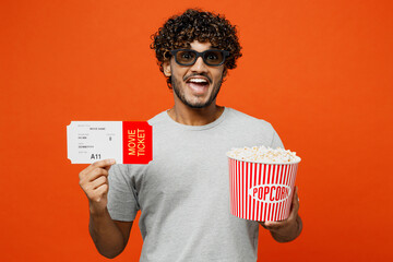 Young smiling shocked happy surprised Indian man in 3d glasses watch movie film hold in hand ticket bucket of popcorn in cinema looking camera isolated on plain orange red background studio portrait. - Powered by Adobe
