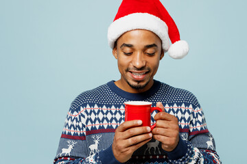 Young satisfied fun man wear knitted sweater Santa hat posing hold in hand cup with coffee or tea isolated on plain pastel blue background. Happy New Year 2024 celebration Christmas holiday concept