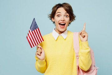 Young shocked woman student wears casual clothes yellow sweater backpack bag hold in hand American flag point index finger up isolated on plain blue background. High school university college concept. - obrazy, fototapety, plakaty