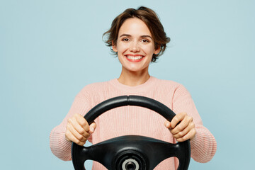 Young smiling happy fun woman wear beige knitted sweater casual clothes hold steering wheel driving car look camera isolated on plain pastel light blue background studio portrait. Lifestyle concept.