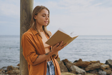 Side view young smart calm student happy woman she wearing orange shirt casual clothes reading book novel study walking on sea ocean sand shore beach outdoor seaside in summer day. Lifestyle concept.