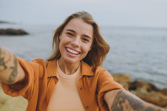 Close up young smiling happy woman she wears orange shirt casual clothes doing selfie shot pov on mobile cell phone walk on sea ocean sand shore beach outdoor seaside in summer day. Lifestyle concept.