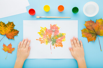 Little child hands showing picture on blue table background. Painting and copying colorful leaf...