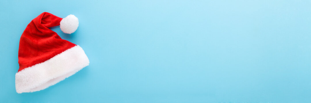 Bright red hat with white fur on light blue table background. Pastel color. Wide banner. Closeup. Garment for Christmas celebration. Empty place for text, quote or sayings. Top down view.