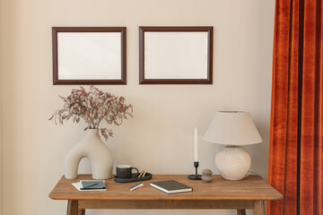 Two empty picture frames on a wall by a table with assorted ornaments, stationary, lighting and a vase filled with flowers