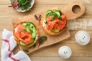Open smoked salmon sandwiches with cream cheese, cucumber, sesame seeds, microgreens, spinach, and peas leaves on light old wooden background. Healthy breakfast food. Delicious snack. Top view.