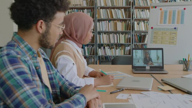 Waist up of multiracial migrant students having online lesson with English teacher via video call on laptop while studying English as foreign language at library