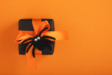 Black gift box with orange ribbon and toy spider on orange background. A place for your text....