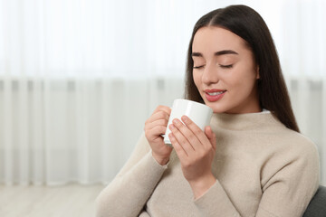 Beautiful young woman holding white ceramic mug at home, space for text