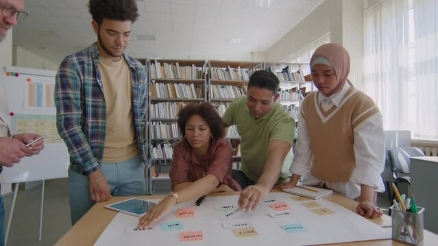 Dolly shot of multiethnic group of migrant students using colorful cards and stickers while playing linguistic game during English class with Caucasian adult male teacher in library